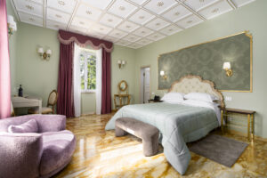 Camera Deluxe Pinewood View di Villa L'Avvenire a Siena: letto con testiera dorata, poltrona rosa e soffitto a rosoni